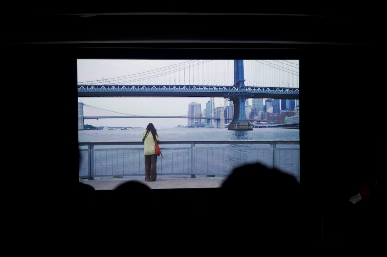 The High School Filmmakers Workshop final presentation attendees sit in a dark theatre-style classroom watching a film project. The still image on the screen shows a young woman with her back to the camera as she looks at the Manhattan Bridge in the daytime.